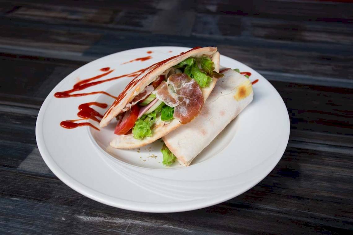 Ein Teller mit einem Sandwich, das mit Salat, Tomaten und Schinken gefüllt ist, und eine rote Sauce.