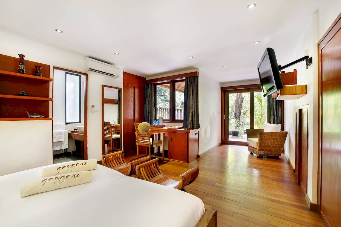 Innenansicht eines Zimmers im Gangehi Island Resort mit Bett, Holzmöbeln, Fernseher und Schreibtisch.