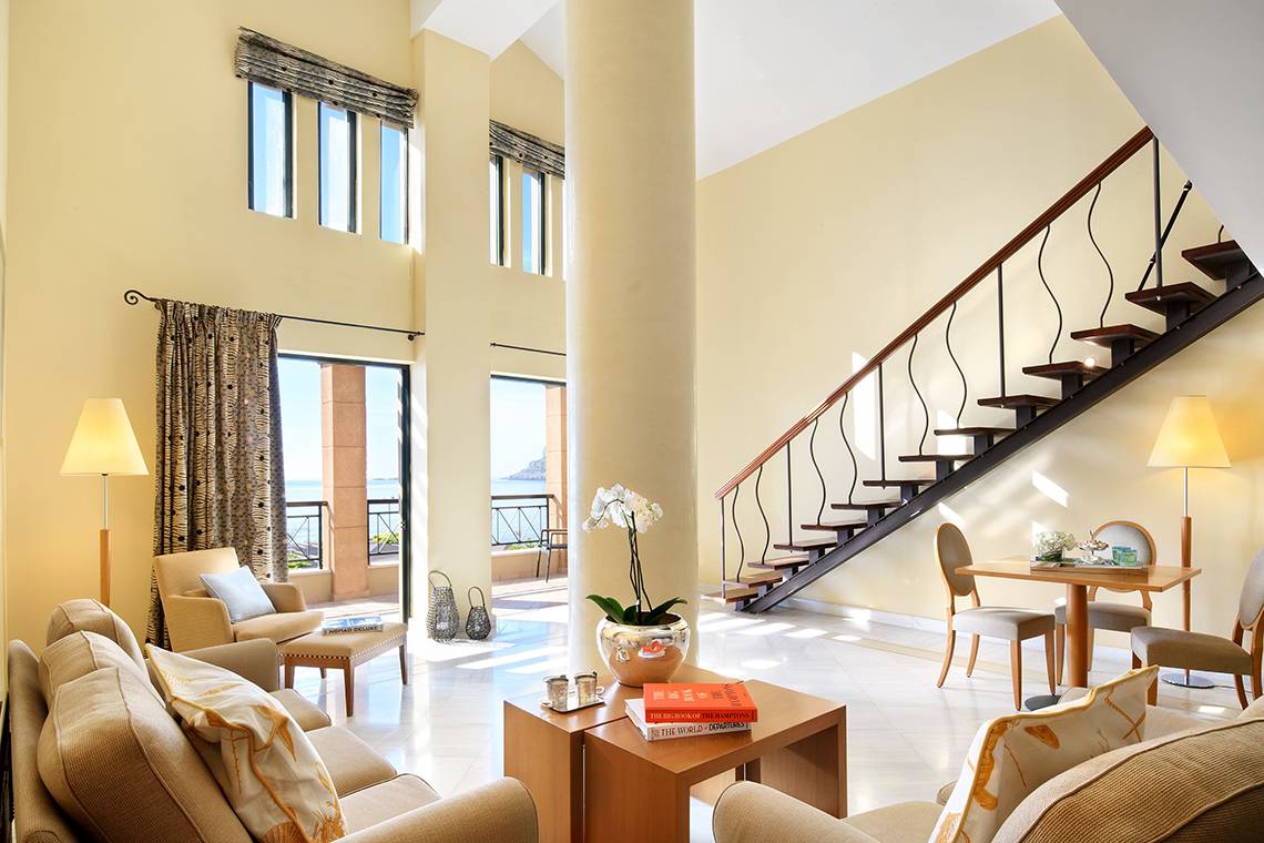 Wohnbereich des Mitsis Lindos Memories Resort & Spa mit Sofas, Tisch, Büchern und Treppe.