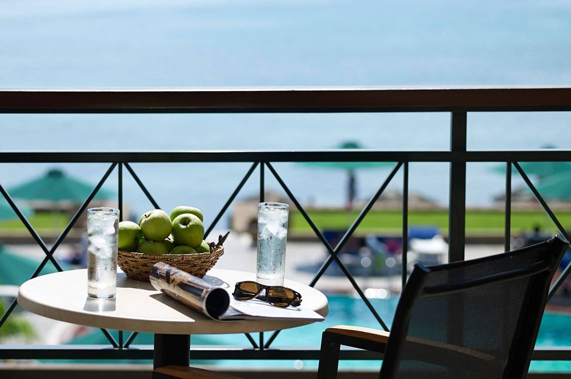 Ein Balkon mit einem Tisch, auf dem eine Zeitschrift, eine Sonnenbrille, ein Glas Wasser und ein Korb mit Äpfeln steht. Im Hintergrund ist ein Pool zu sehen.