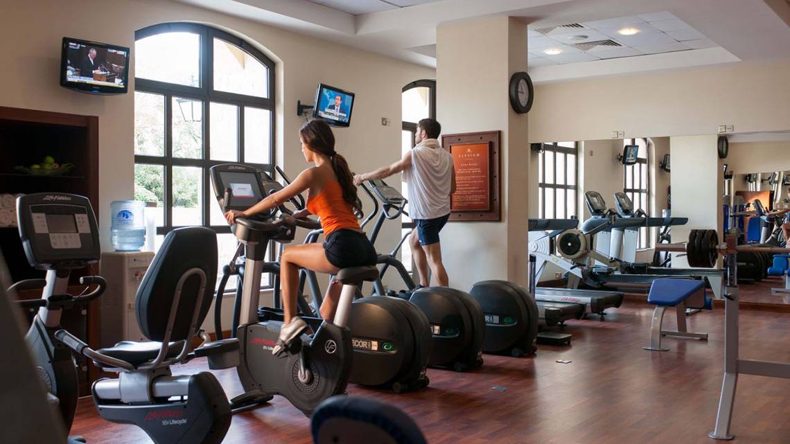 Eine Frau und ein Mann trainieren im Fitnessraum des Elysium Hotels auf einem Ellipsentrainer.