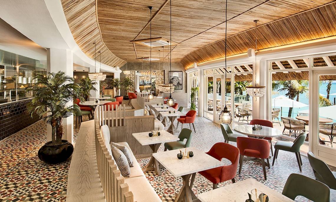 Innenansicht des Restaurants im LUX Grand Gaube Resort & Villas mit bunten Stühlen und Tischen, bepflanzten Ecken und Blick auf den Ozean.
