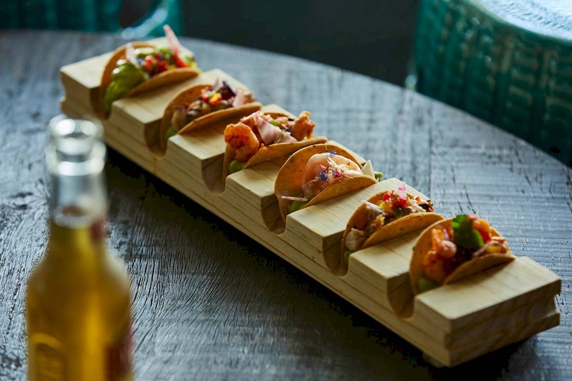 Ein Holzbrett mit verschiedenen gefüllten mexikanischen Tacos auf einem Tisch.