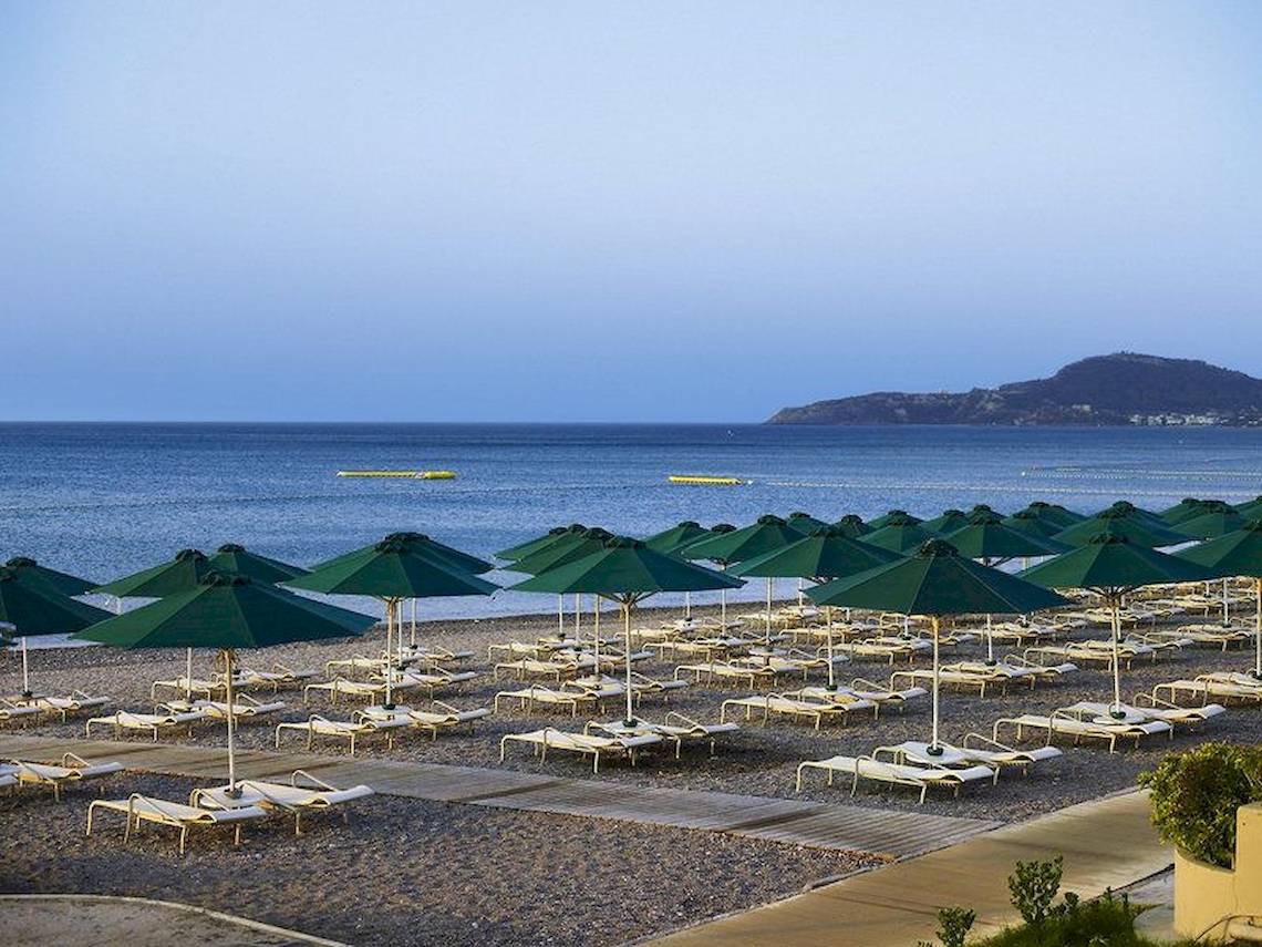 Foto der Strandseite des Esperos Village Blue & Spa Hotels mit vielen Sonnenliegen und grünen Sonnenschirmen.