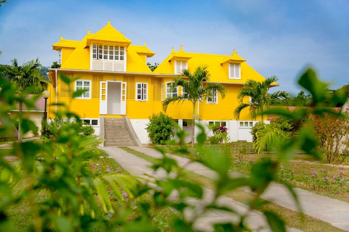 Außenansicht des gelben La Digue Island Lodge Hotels mit Gehweg und Palmen.