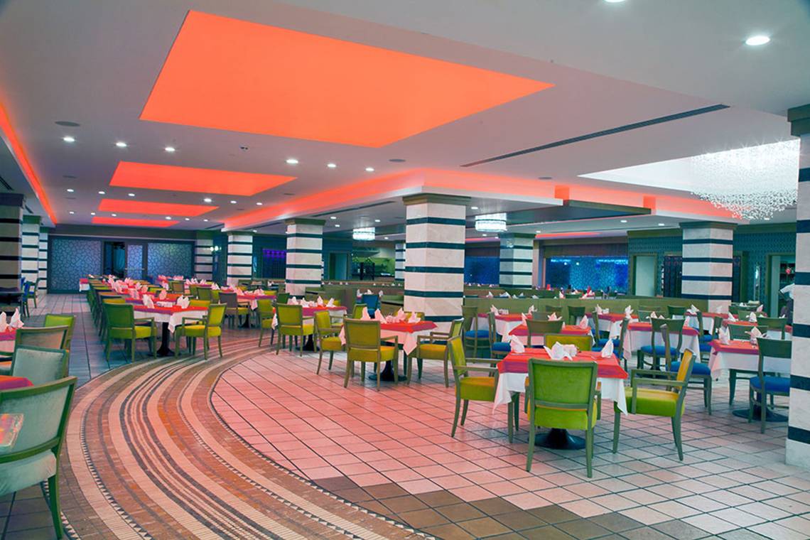 Innenansicht des Restaurants im Belconti Resort mit runden Tischen, farbigen Stühlen und orangefarbenen Deckenleuchten.