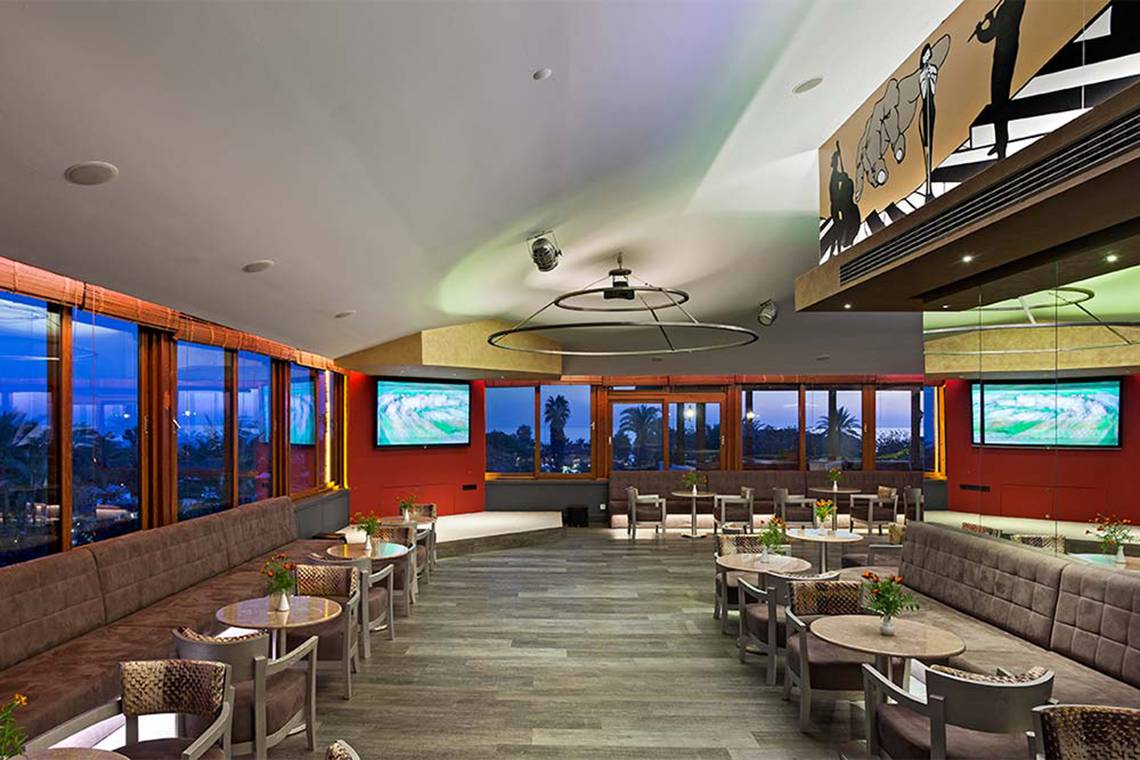 Innenansicht der Lounge im Belconti Resort mit Sitzbereichen, Tischen und Fernsehern.