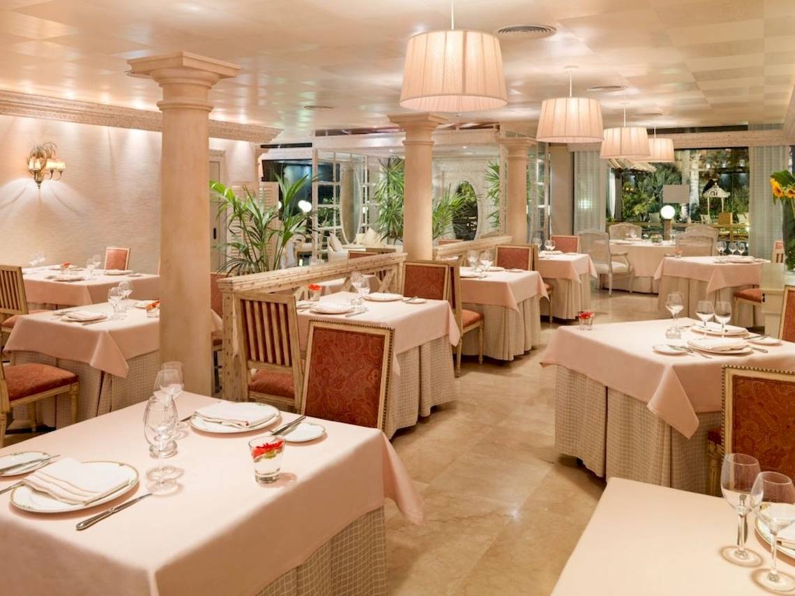 Innenansicht des Restaurants des Secrets Bahia Real Resort & Spa mit gedeckten Tischen, eleganten Stühlen und beleuchteten Kronleuchtern.