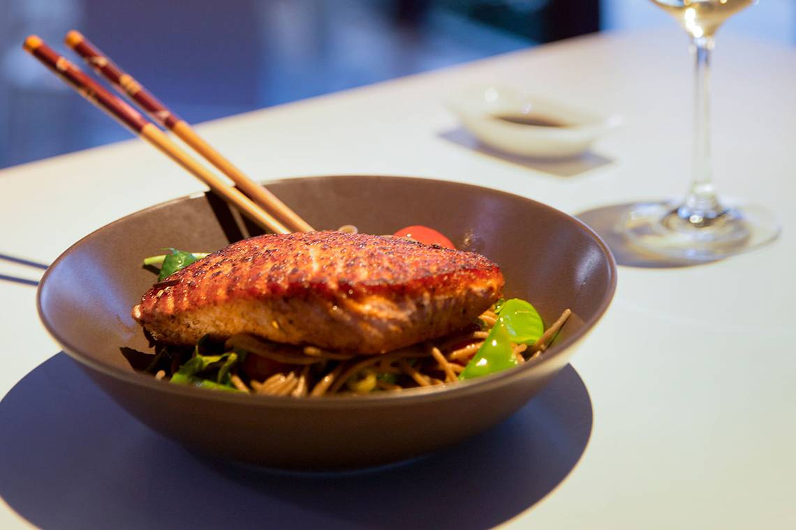 Gebratener Lachs in einer Schüssel mit Nudeln und Gemüse, serviert mit Chopsticks und einem Glas Wein.