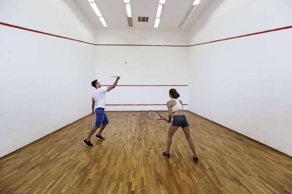 Zwei Personen spielen Squash in einer Sporthalle mit Holzboden und roten Linien.