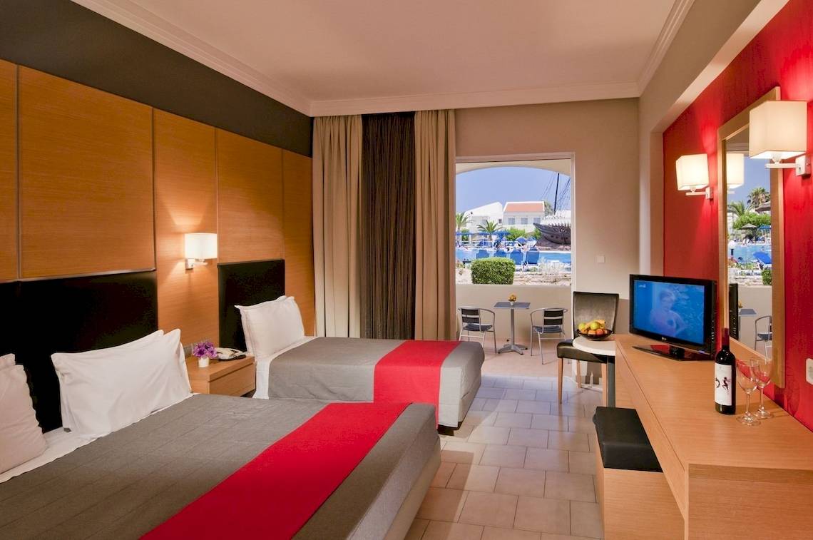 Innenansicht eines Doppelzimmers im Kipriotis Village Resort mit zwei Betten, Fernseher und Balkonsicht.