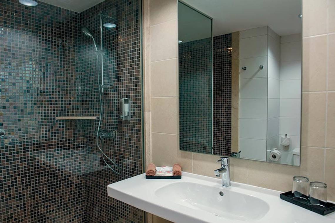 Badezimmer im Riu Lupita Hotel mit gefliester Wand, Dusche, Waschbecken und Spiegel.