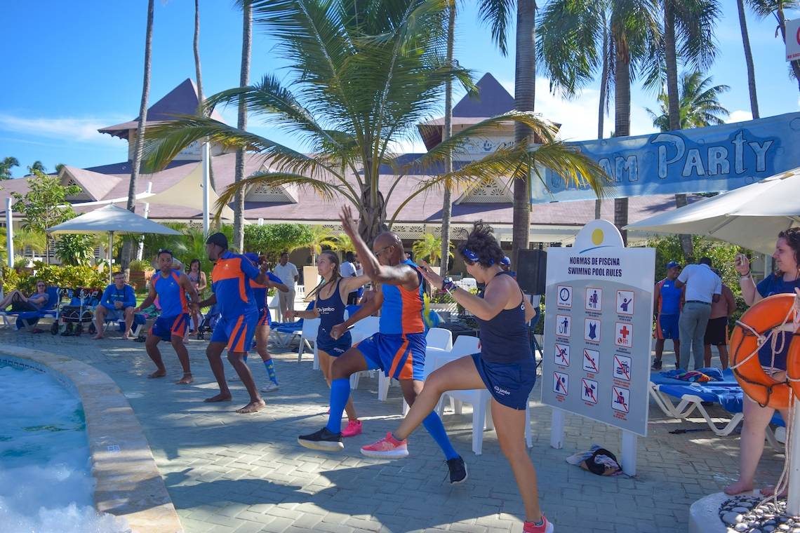 Gruppe von Personen in blauen und orangefarbenen Trainingsanzüge beim Tanzen am Pool im Vista Sol Punta Cana Beach Resort & Spa.