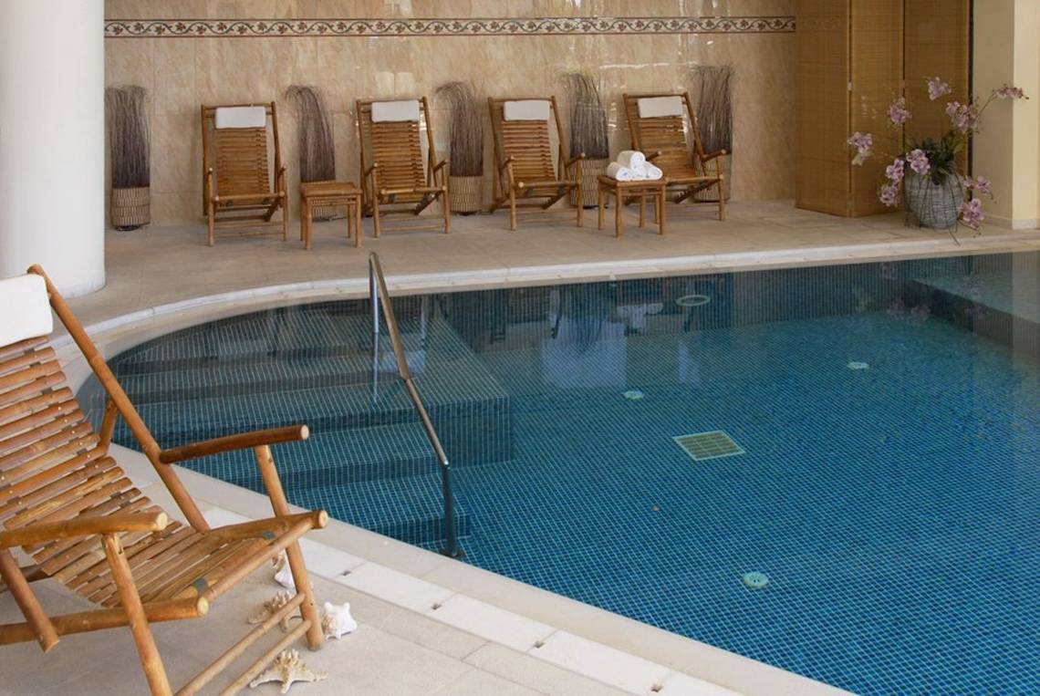 Innenpool des Aquila Porto Rethymno Hotels mit Holzstühlen und Handläufen.