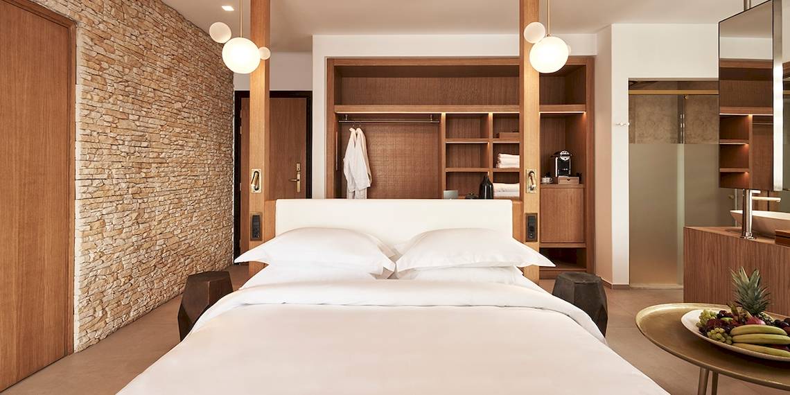 Schlafzimmer im Myconian Naia Hotel mit Steinwand, Holzschrank und frischem Obst auf dem Tisch.