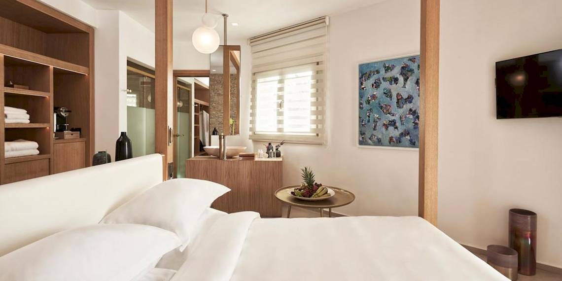 Schlafzimmer im Myconian Naia Hotel mit weißem Bett, weißem Kopfteil, Nachttisch, Fernseher an der Wand und Badezimmer im Hintergrund.