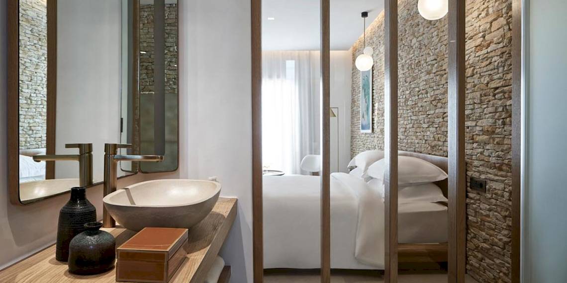 Schlafzimmer im Myconian Naia Hotel mit einem Bett, Kissen, einem Steinwand, einem Waschbecken, Spiegel und Vasen.