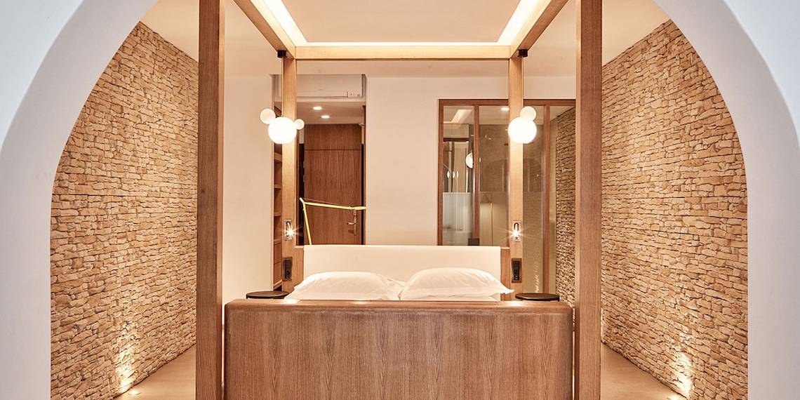 Schlafzimmer im Myconian Naia Hotel mit Holzbett und weißen Kissen, umgeben von Steinwänden.