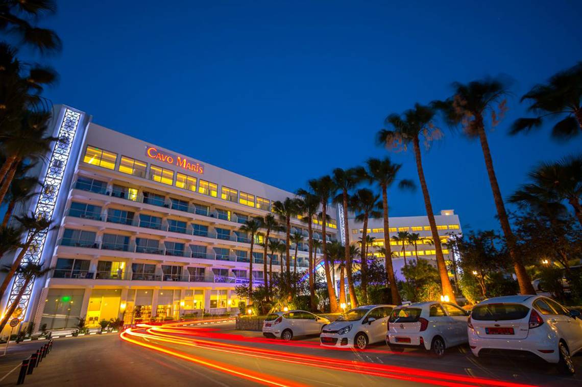 Cavo Maris Beach Hotel bei Nacht mit beleuchteten Zimmern und Autos vor dem Gebäude.