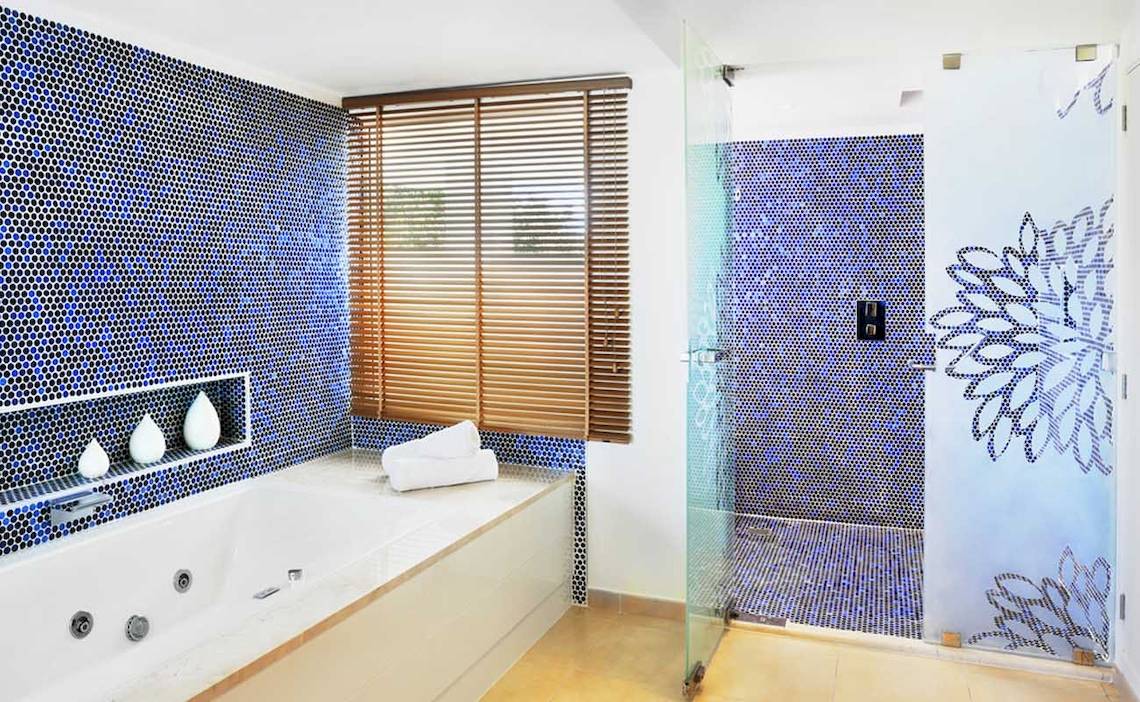Ein Badezimmer mit blauen Fliesen, Badewanne, Dusche und Fenster mit Jalousien.