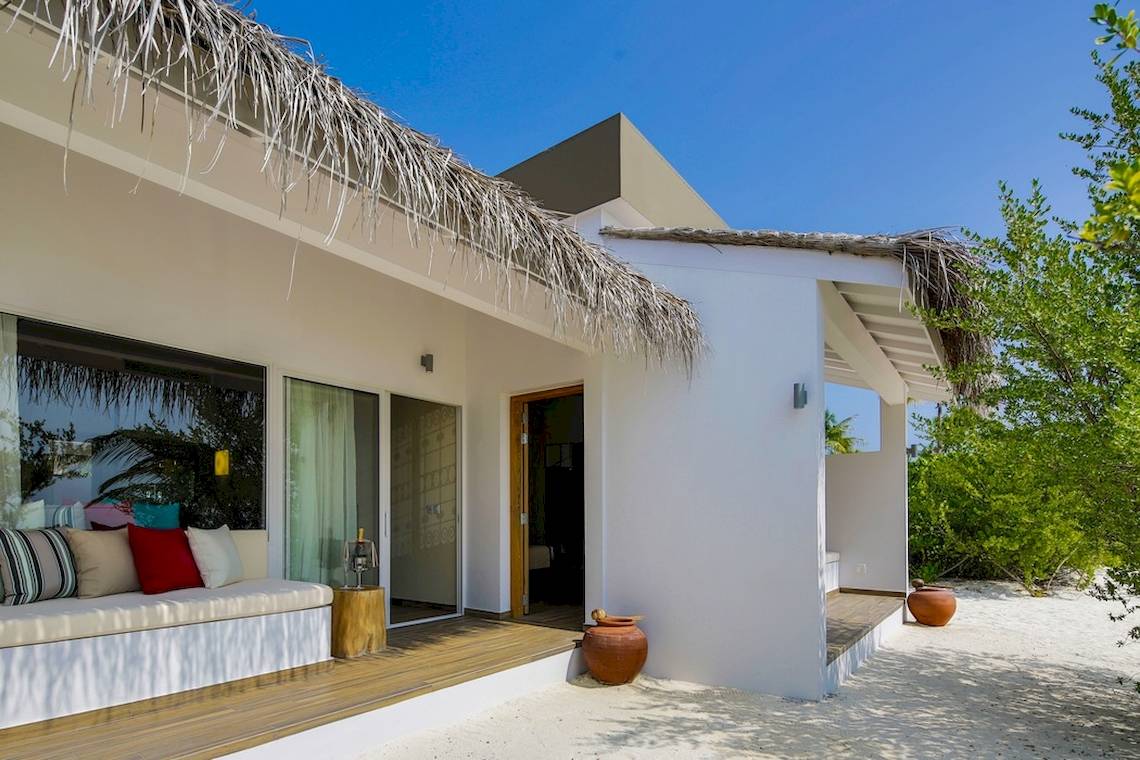 Außenbereich der Cocoon Maldives Villa mit einem weißen Gebäude, einem Holzsofa, großen Fenstern und einer Strohdach.