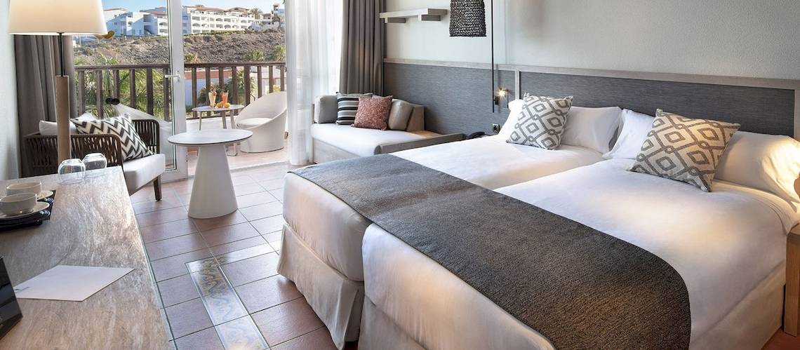 Doppelzimmer im Fuerteventura Princess Hotel mit Balkon, zwei Betten, Tisch und Aussicht auf die Stadt.