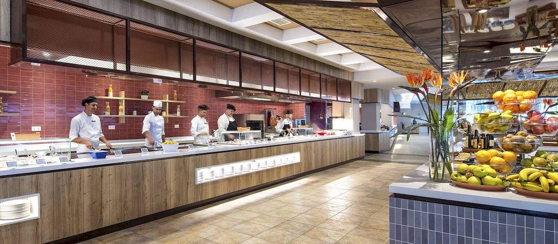 Innenansicht des Restaurants im Fuerteventura Princess Hotel mit Köchen, die an einer langen Theke arbeiten und verschiedene Früchte auf dem Tisch sind.