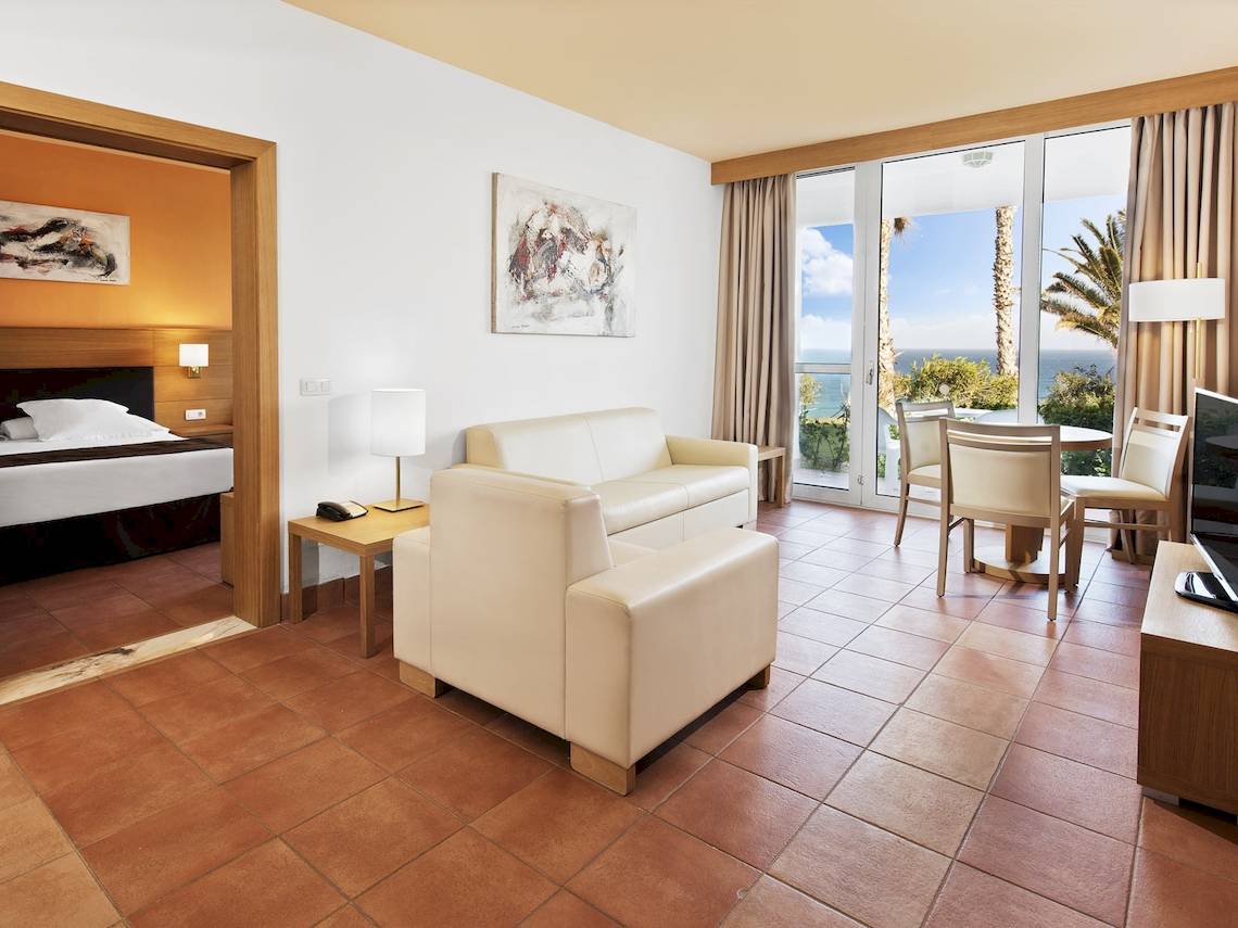 Innenansicht eines Zimmers im Fuerteventura Princess Hotel mit Bett, Wohnzimmerbereich, Esstisch und Meerblick.
