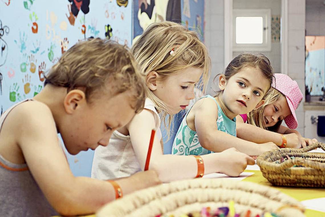 Vier Kinder sitzen an einem Tisch und zeichnen, ein Korb mit Buntstiften steht vor ihnen.