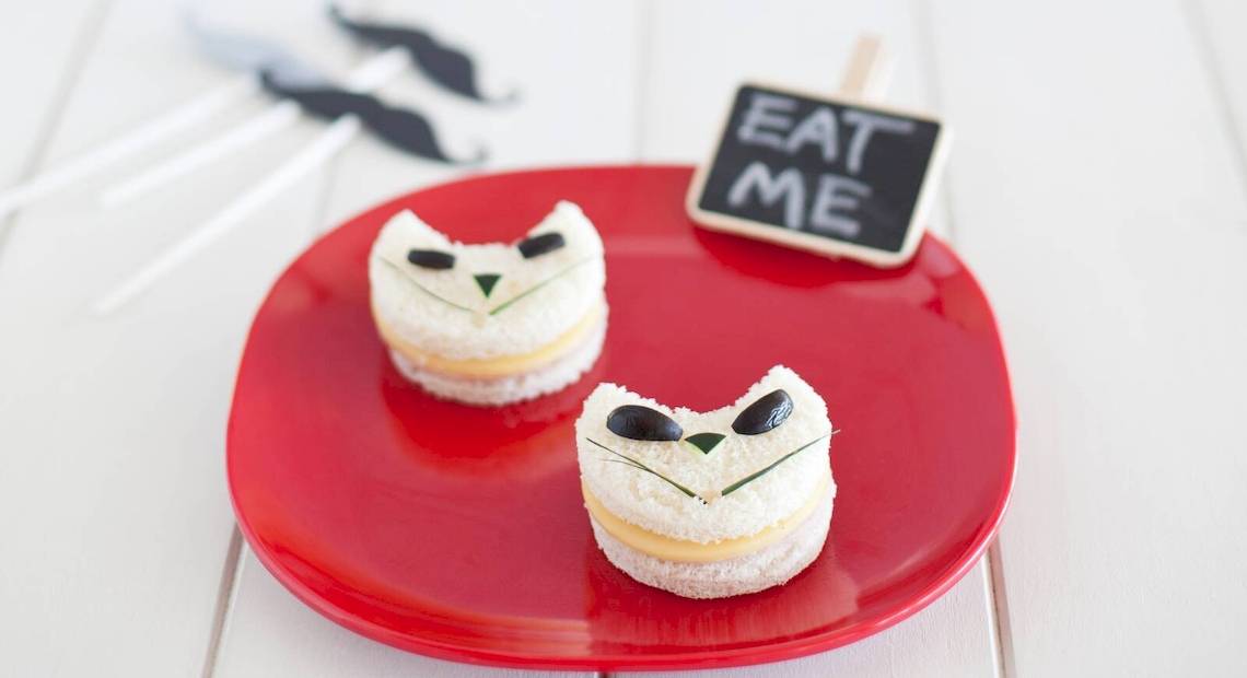 Zwei Katzen-Sandwiches auf einem roten Teller mit einem schwarzen Schild, das 'Eat Me' sagt, und Mustache-Picks im Hintergrund.