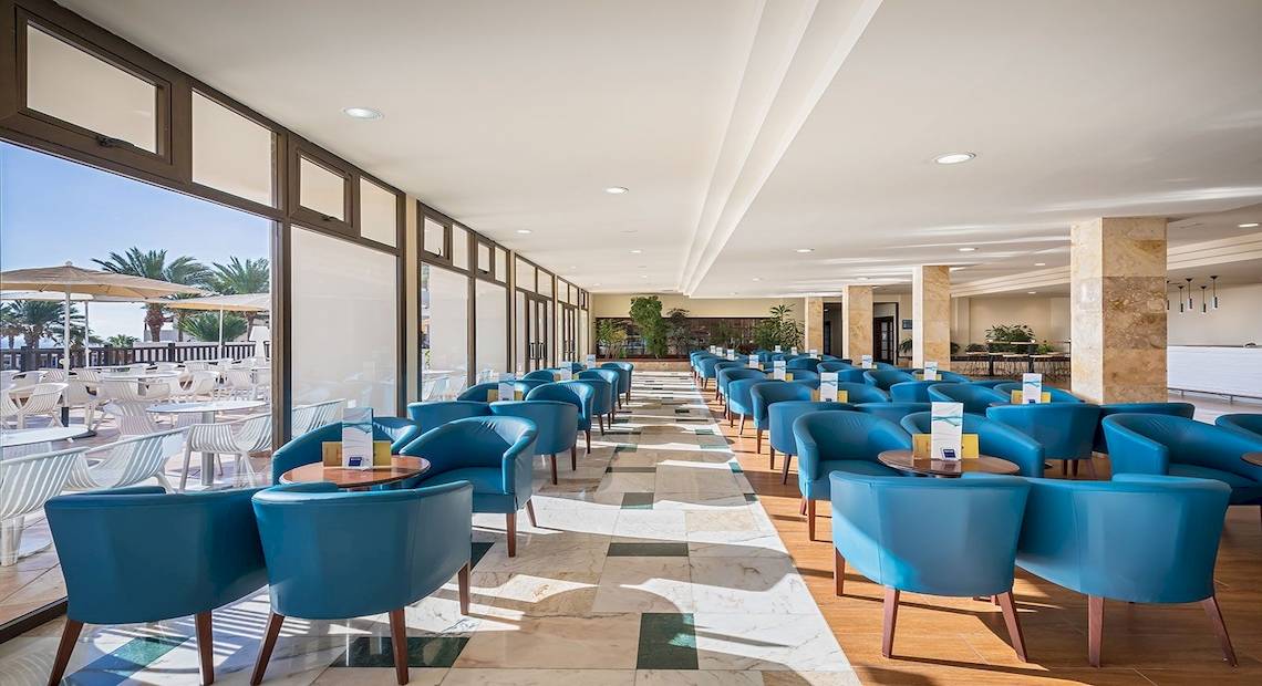 Innenbereich des Occidental Jandia Mar Hotels mit vielen blauen Stühlen und Tischen, große Fenster und Sonnenschirme draußen