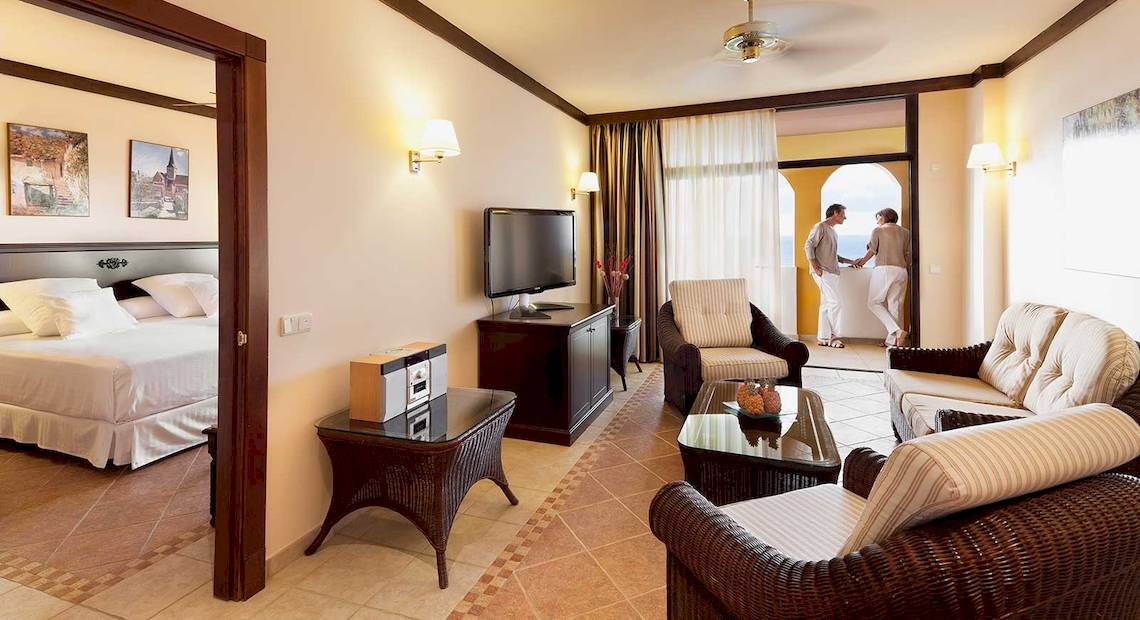 Innenansicht einer Suite im Occidental Jandia Royal Level Hotel mit Schlafzimmer, Wohnzimmer und Balkon.