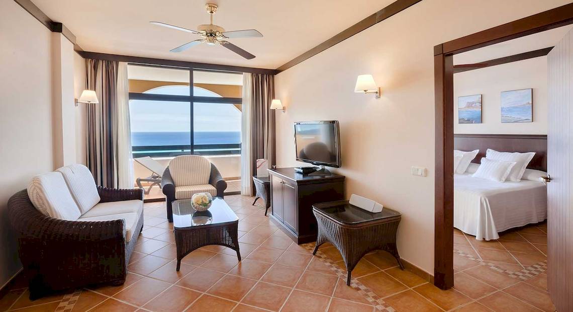 Hotelzimmer mit Meerblick, Wohnzimmerbereich mit Sofa, Tisch, Fernseher und Balkon.