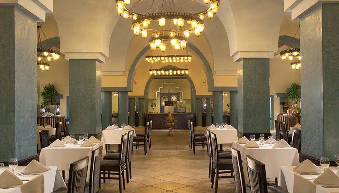Innenansicht des Restaurants im Jaz Lamaya Resort mit gedeckten Tischen, Kronleuchtern und blauen Säulen.