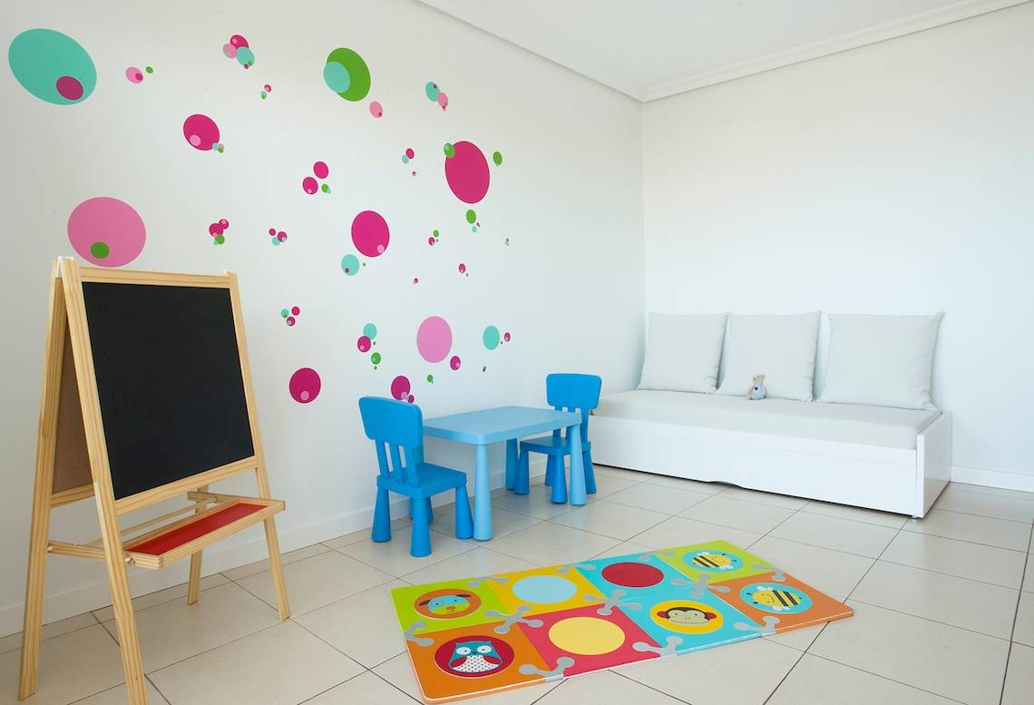 Ein Kinderzimmer mit einem blauen Spieltisch, zwei blauen Stühlen, einer Bank, einem Teppich und einer Tafel.