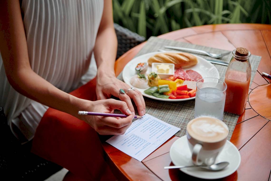 Frau schreibt auf einem Papier, während sie ein Frühstück genießt, bestehend aus Gemüse, Fleisch und Brot, im Salinda Resort auf Phu Quoc Island.