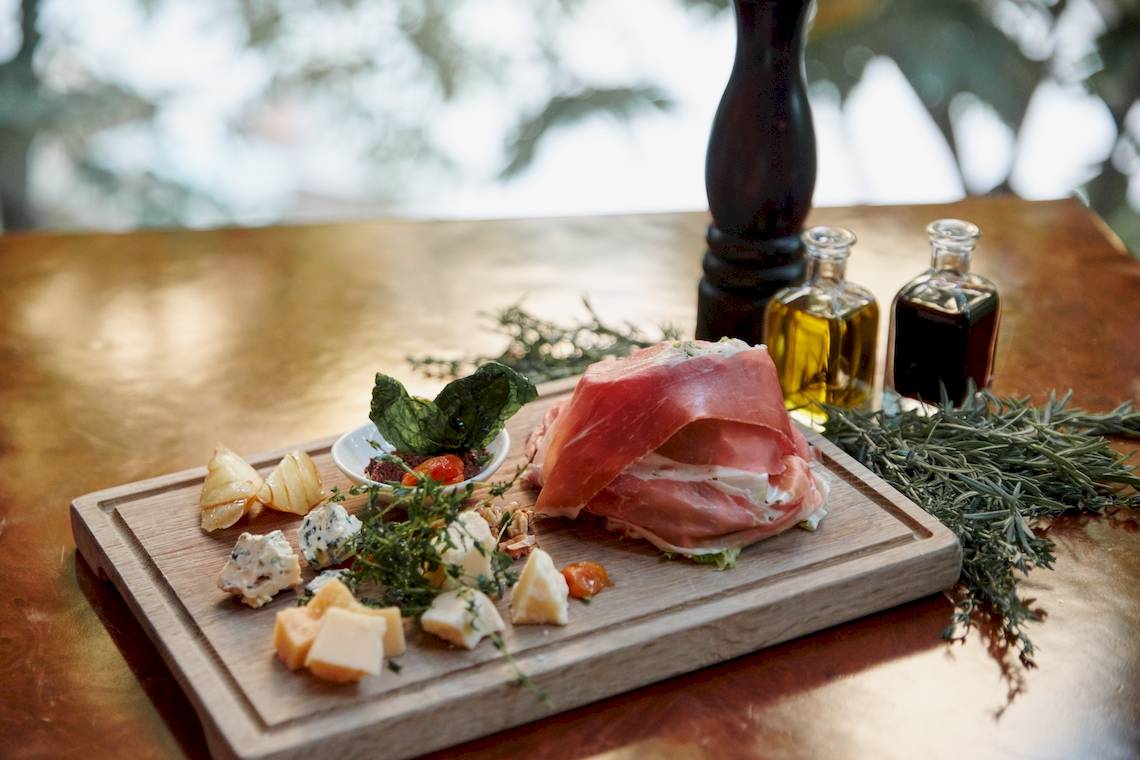 Ein Holzschneidebrett mit Prosciutto, verschiedenen Käsesorten und Kräutern auf einem Tisch.