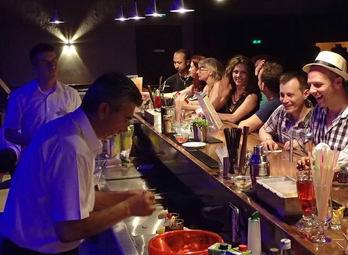 Ein Barkeeper mischt Getränke in einer Hotelbar mit blauer Beleuchtung, während Gäste an der Theke sitzen.
