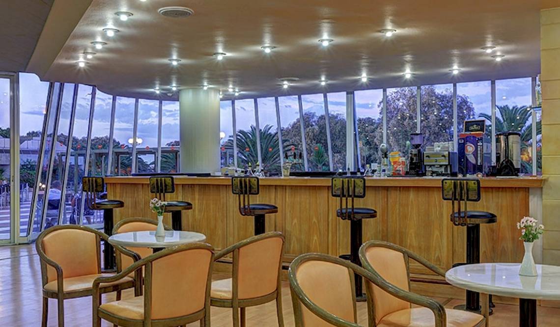 Innenansicht der Bar im Giannoulis Santa Marina Beach Hotel mit runden Tischen, Stühlen und einer Theke mit Getränken.