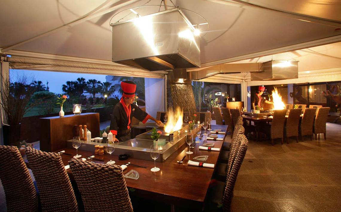 Ein Koch bereitet am Tisch im GrandResort Limassol ein Gericht mit einem Feuergrill zu.