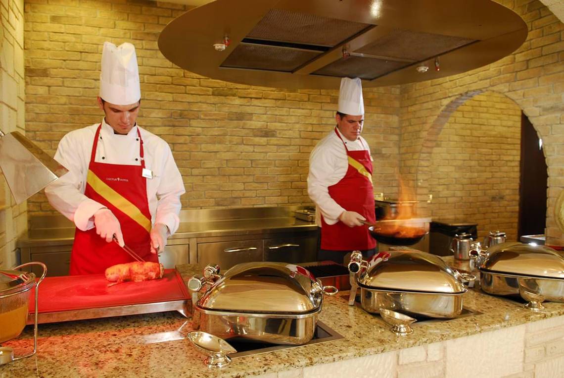 Zwei Küchenchefs im Cactus Royal Hotel bereiten Speisen zu, einer schneidet ein Gericht auf einem Schneidebrett, der andere brät etwas in einer Pfanne.
