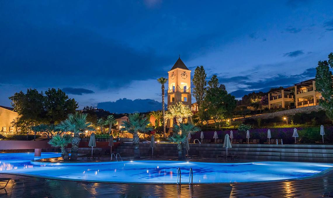 Candia Park Village Hotel Außenbereich mit beleuchtetem Pool und Uhrturm bei Nacht
