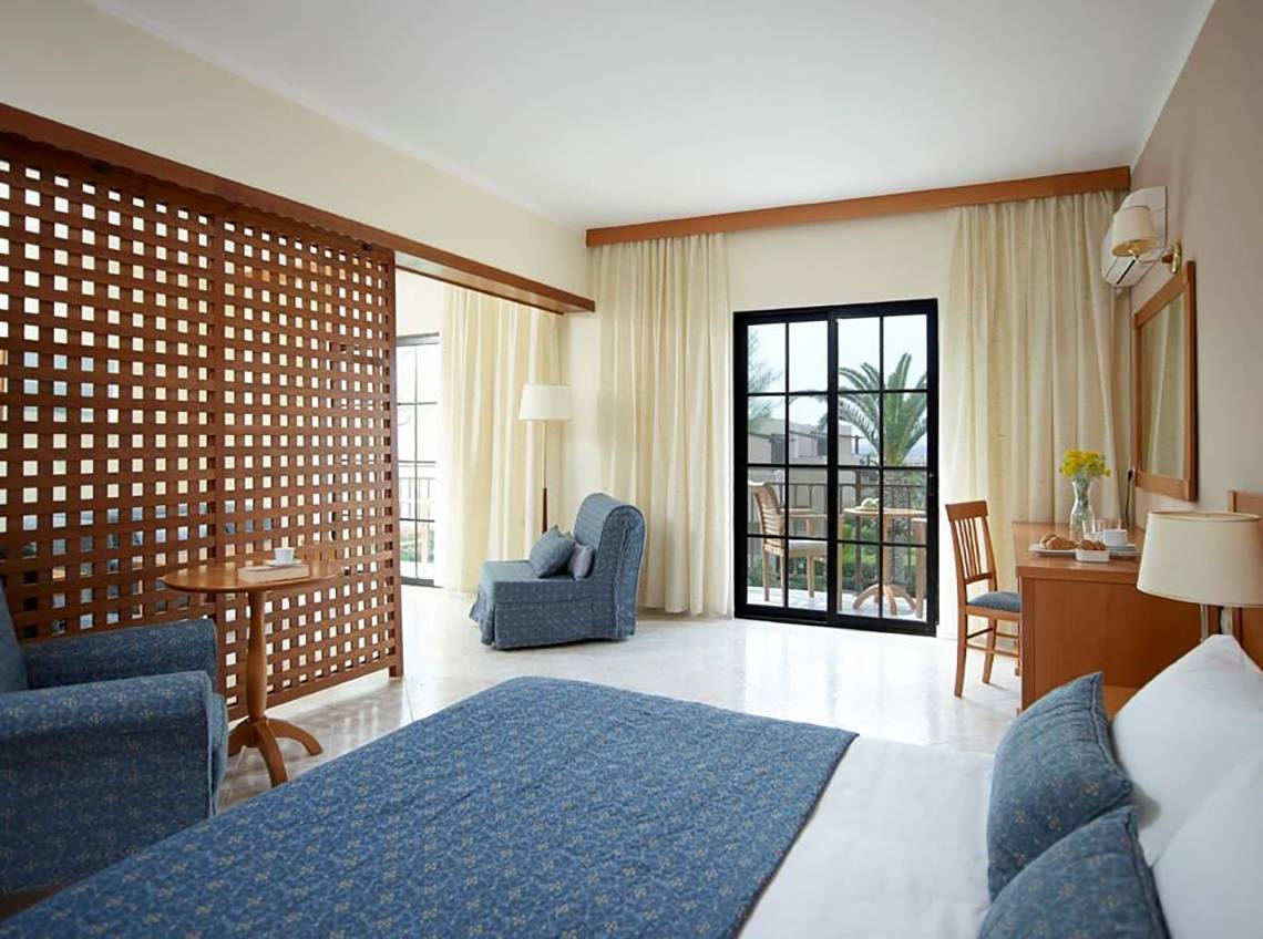 Innenansicht eines Zimmers im Porto Bello Beach Hotel mit Bett, Sofa, Tisch und Terrassentür.