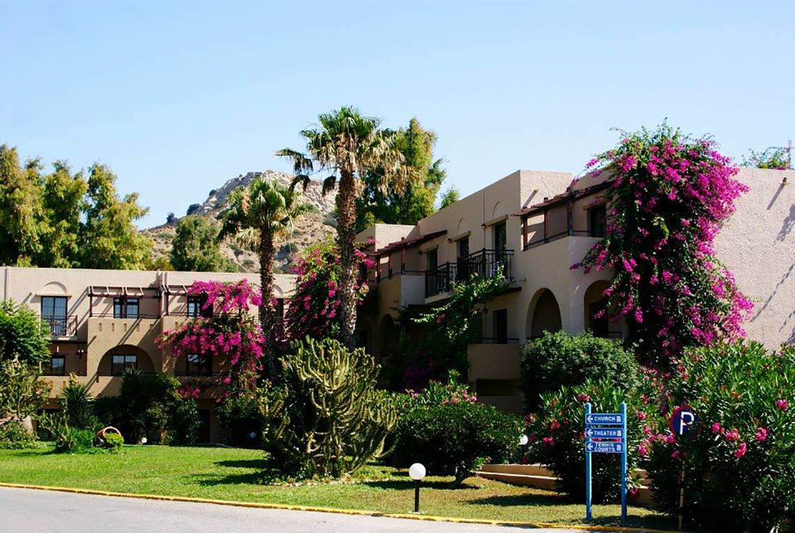 Außenansicht des Porto Bello Beach Hotels mit grüner Rasenfläche, Palmen und Blumenbüschen.