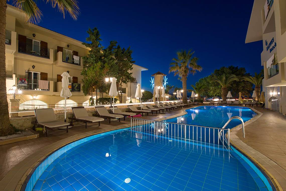 Pool-Bereich des Kalyves Beach Hotels bei Nacht mit Lounge-Liegen und Gebäude im Hintergrund.