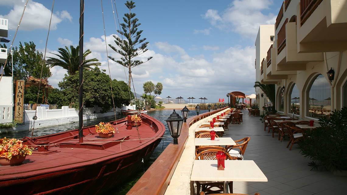 Terrasse des Kalyves Beach Hotels mit einem roten Boot im Kanal