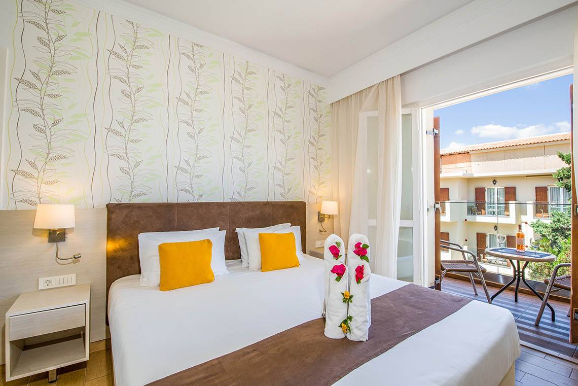 Zimmer im Kalyves Beach Hotel mit Balkon, Bett, Nachttischen und Ausblick auf eine Terrasse.