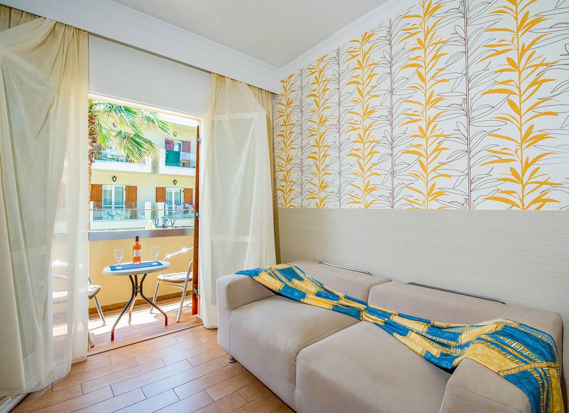 Zimmer im Kalyves Beach Hotel mit Balkon, Sofa, Tisch und Blick auf die Straße.