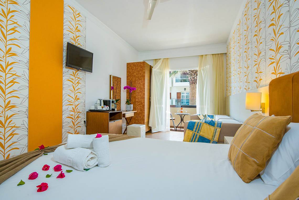 Innenansicht eines Zimmers im Kalyves Beach Hotel mit farbenfrohen Wänden, einem Bett, Fernseher und Balkon.
