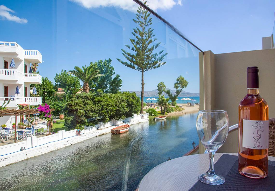 Aussicht von Kalyves Beach Hotel auf Strand und Kanal mit einem Glas und einer Flasche Wein auf dem Tisch.